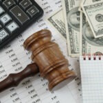 a calculator, gavel, hundred dollar bills, and a notepad on top of a printed spreadsheet