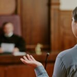 Woman speaking at witness stand with microphone before judge in courtroom