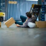 Warehouse worker lying on floor beside hard hat and fallen cardboard boxes