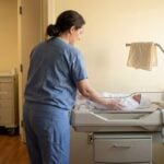 nurse in scrubs tending to swaddled newborn in hospital bassinet