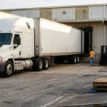 semi-truck backing into warehouse loading dock with workers in safety vests nearby